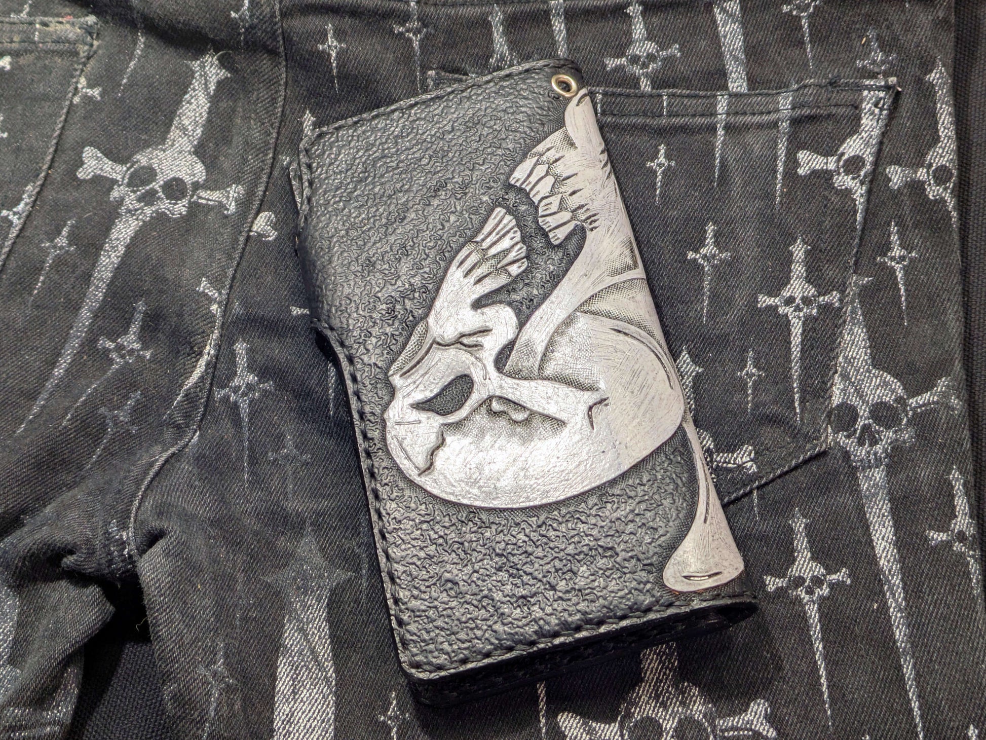 Back view of the black leather wallet with a white skull design, mirroring the front. Textured embossing and side stitching detail visible on a non-specific textured background created by a tooling mark.