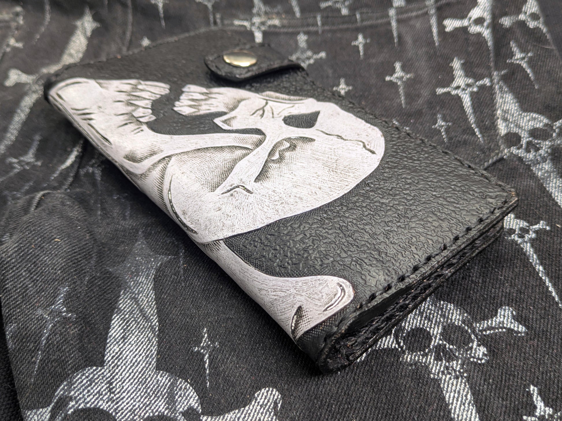 Black leather wallet with white double skulls Ace of Spades design, a slightly varied frontal perspective. Embossed texture and visible snap button fastener.