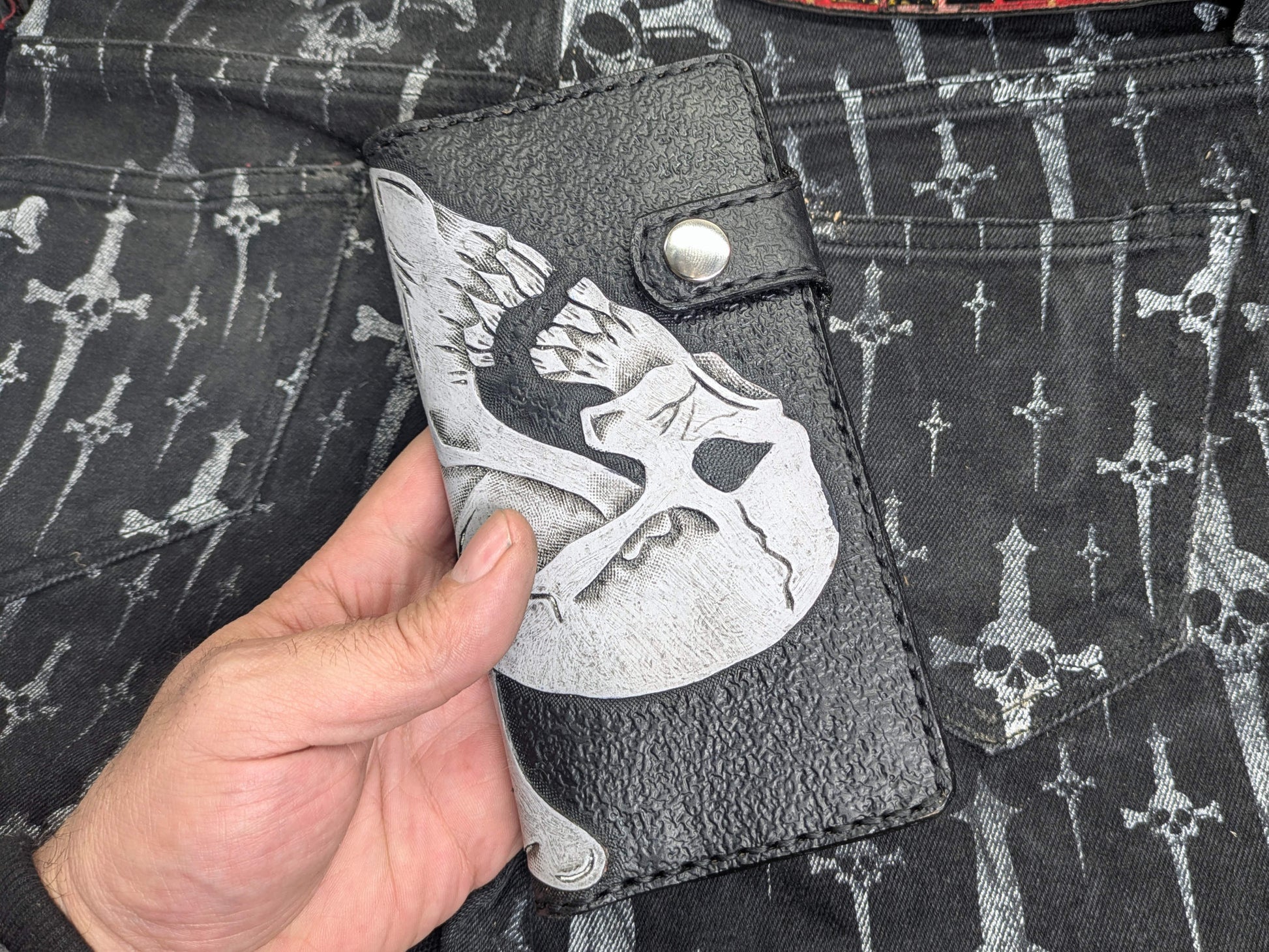 Hand holding black leather wallet with white double skulls Ace of Spades design. Front view shows textured leather and snap button closure, providing a size perspective.