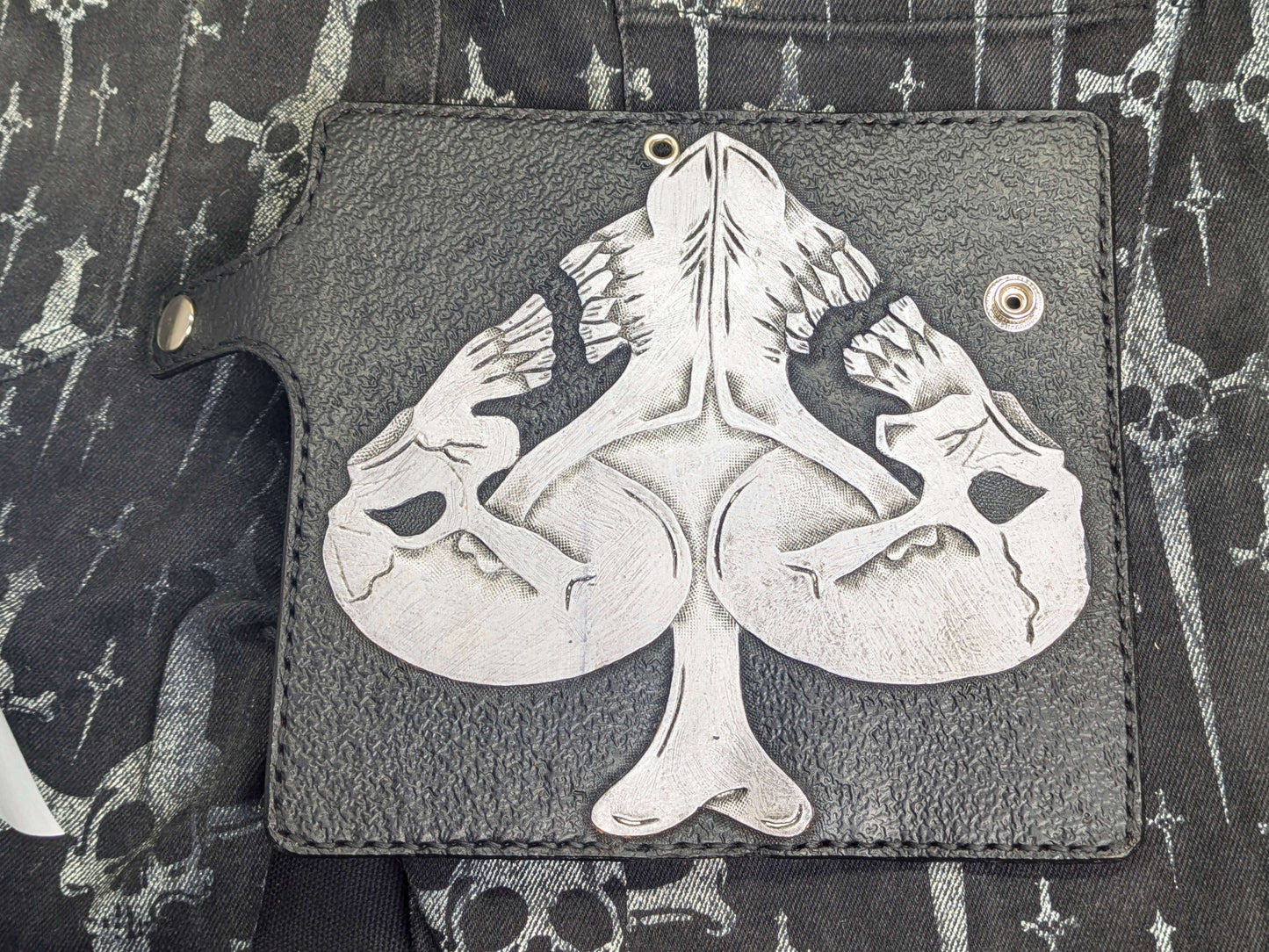 Open view of the black leather wallet, showcasing the full Ace of Spades design composed of two white skulls and a central bone. Visible snap button closure, leather strap, and metal eyelet for chain attachment.