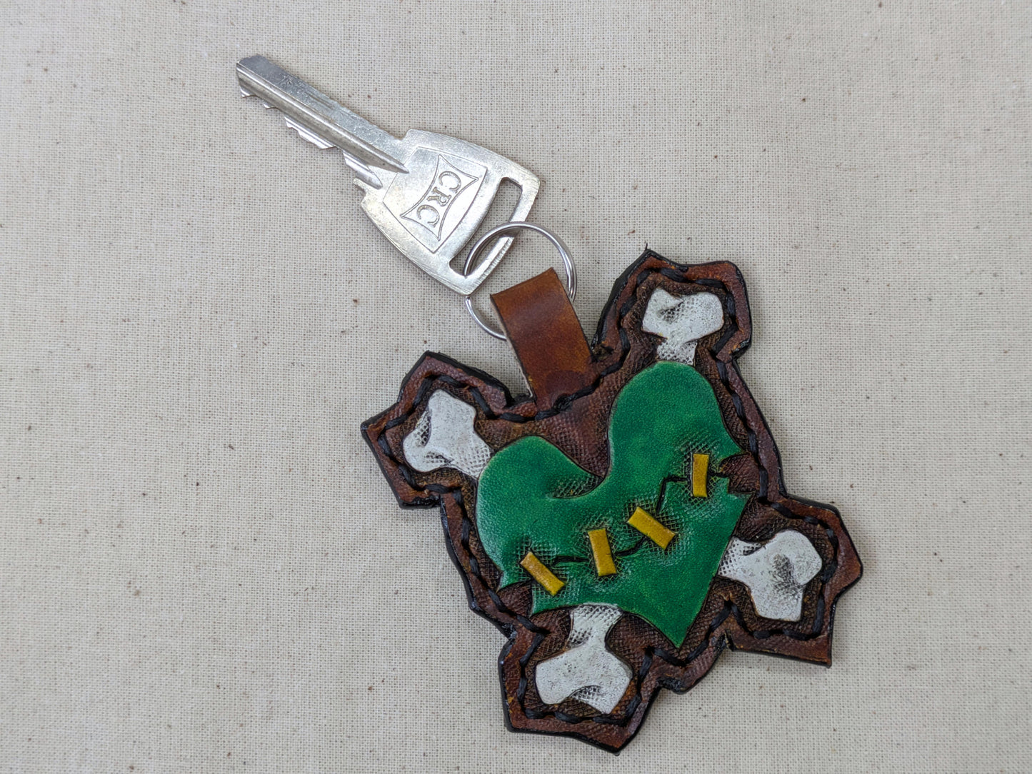 Handmade leather zombie heart keychain, green with white bones and yellow stitches, attached to a car key, on a light fabric surface.