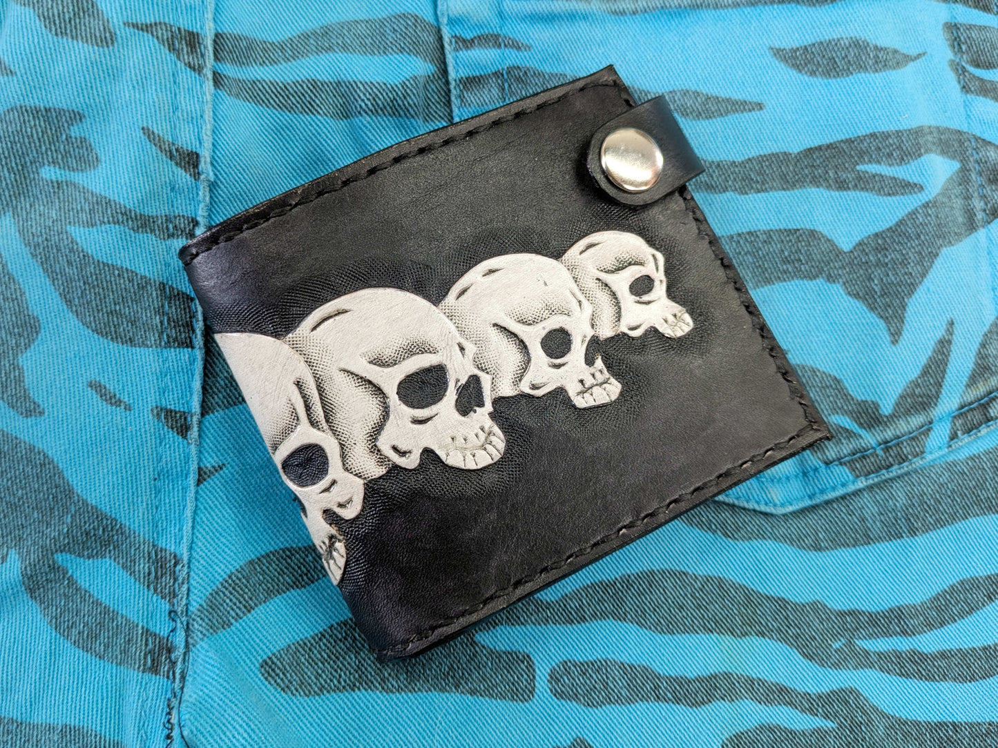 Handmade black leather bifold wallet with three white skulls embossed in relief on the front, closed with a snap button, on a blue zebra-patterned fabric.