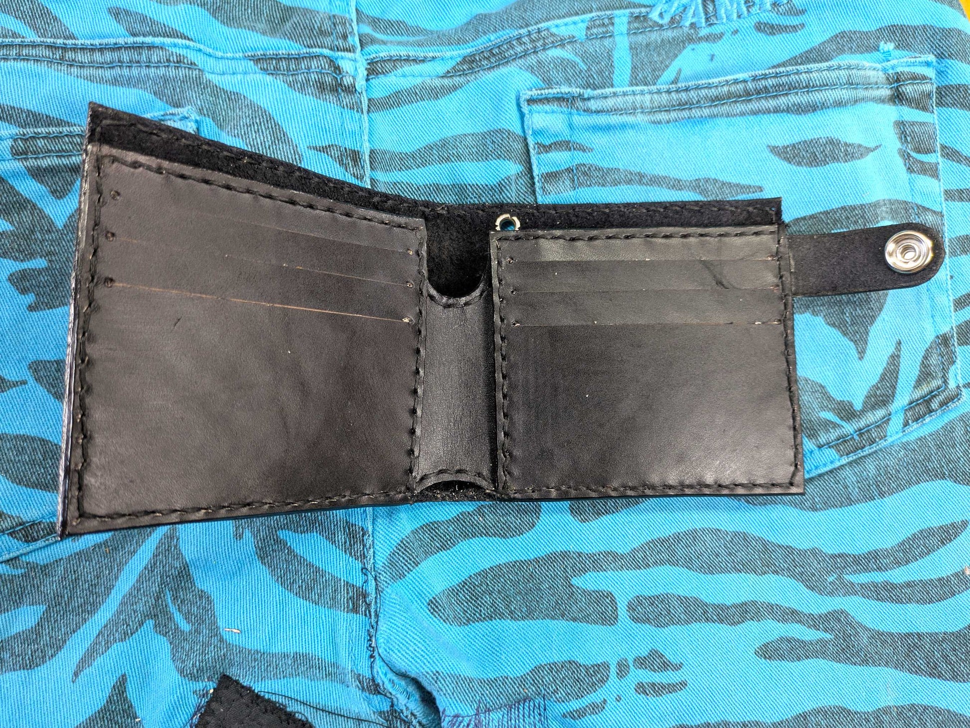 Open black leather bifold wallet, showing the interior with six card slots and a note compartment, with black hand-stitching, on a blue zebra-patterned fabric.