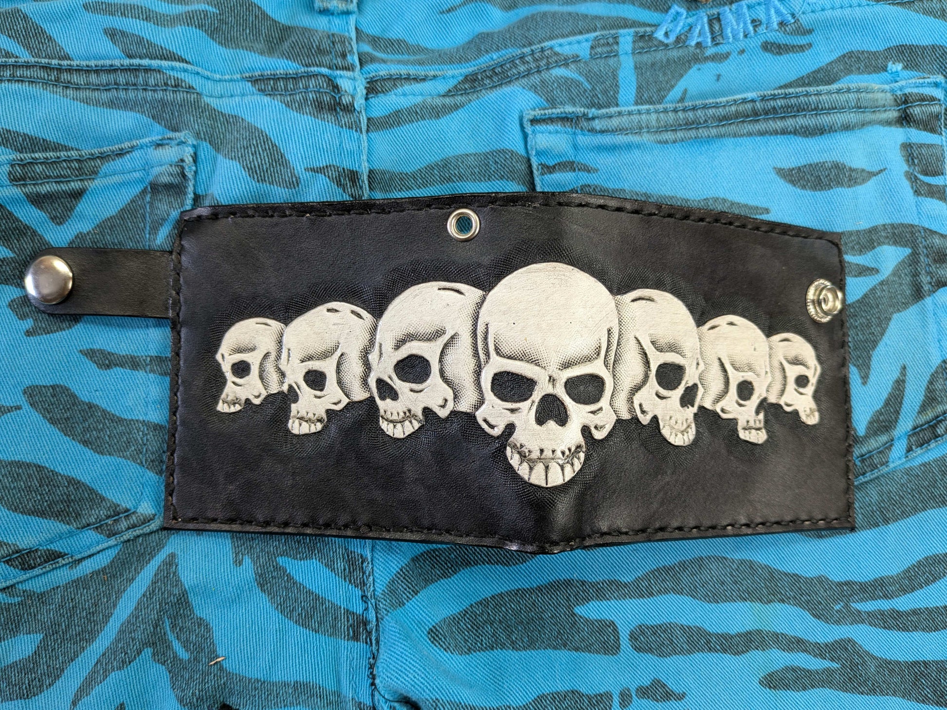 Back view of the black leather bifold wallet featuring a row of seven white skulls embossed in relief, with a snap-button closure tab, on a blue zebra-patterned fabric.