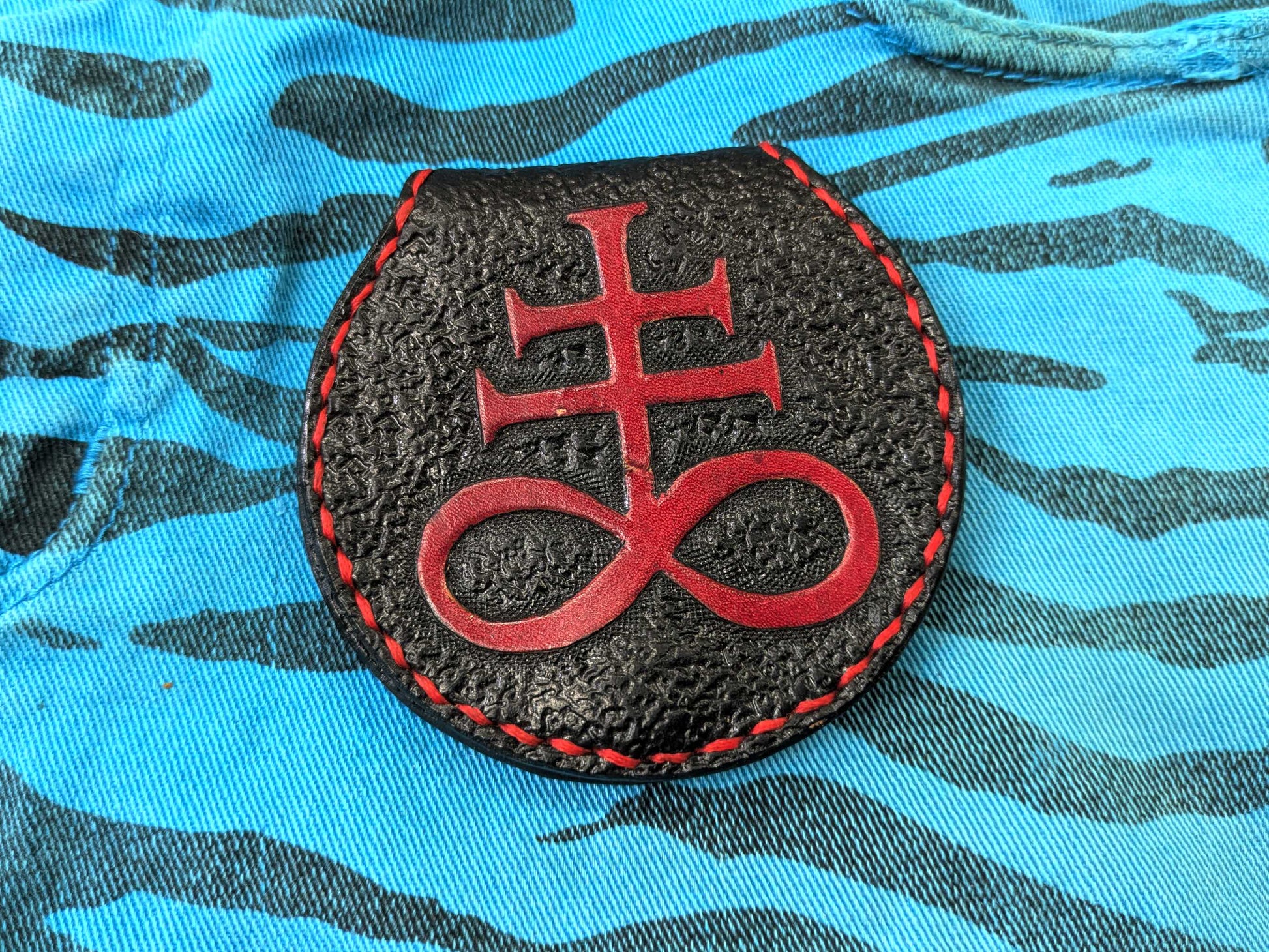 Handmade black leather coin purse with an engraved red Leviathan Cross, featuring a textured pattern and red stitching, on a blue zebra-patterned fabric.
