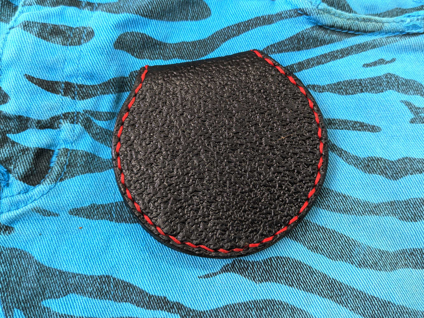 Back view of the black leather Leviathan Cross coin purse, showing the uniform textured pattern and red hand-stitching, on a blue zebra-patterned fabric.