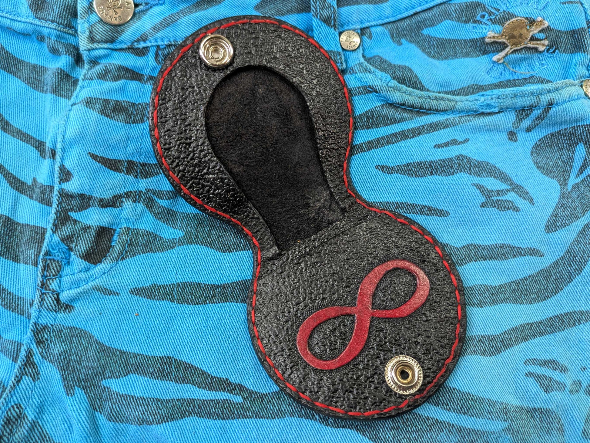 Open interior of the black leather Leviathan Cross coin purse, revealing the engraved red Infinity Symbol, rough texture, red stitching, and compartments for coins and folded notes, on a blue zebra-patterned fabric.