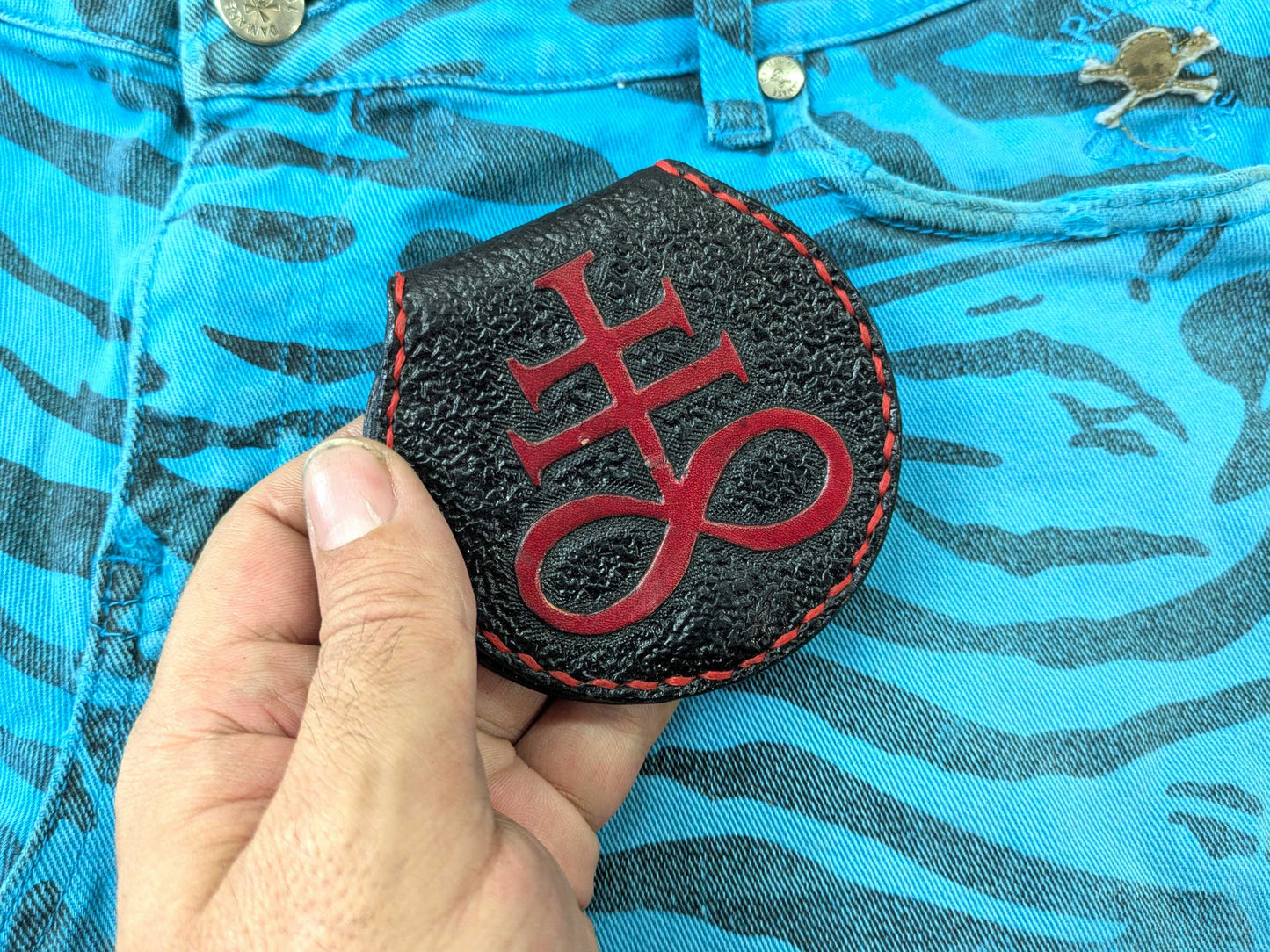Hand holding the black leather coin purse with the engraved red Leviathan Cross, displaying its texture and stitching, against a blue zebra-patterned fabric.