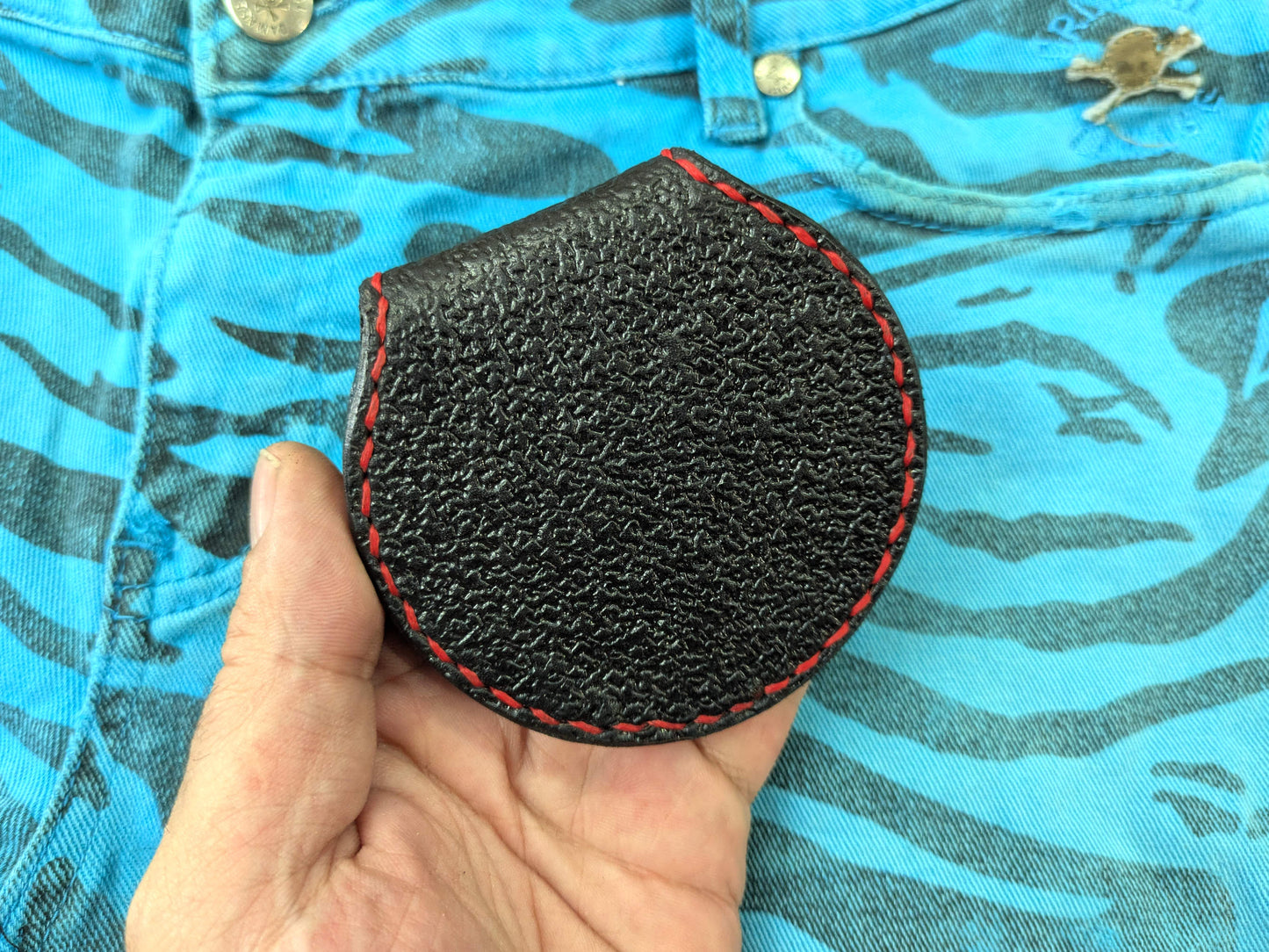 Hand holding the back of the black leather Leviathan Cross coin purse, displaying its uniform textured pattern and red hand-stitching, against a blue zebra-patterned fabric.
