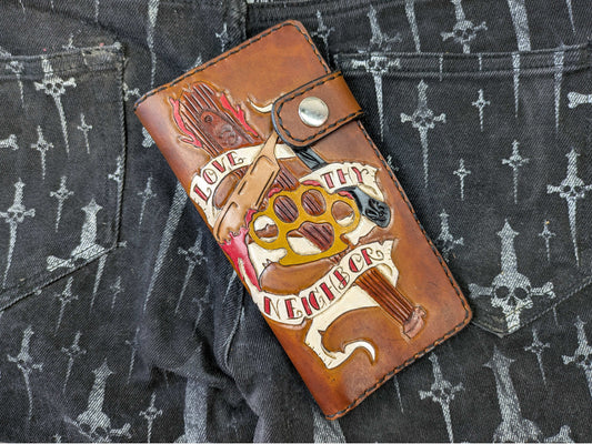 Brown leather wallet with "Love Thy Neighbor" engraved design, featuring brass knuckles, knife, and torch, viewed from above on a dark surface with a cross/skull pattern.